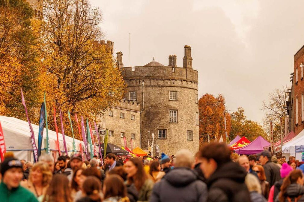 A crowded outdoor street scene in front of a stone castle, with autumn trees, colorful event tents, and many people in the foreground under an overcast sky.